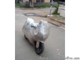 The image shows a motor scooter parked on the side of a street. The scooter is covered with a transparent plastic cover, whic...