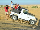The image shows a group of five people standing in front of a white jeep in the desert. The jeep is parked on a sandy dune wi...