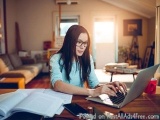 The image shows a young woman sitting at a wooden desk in a living room. She is wearing a blue shirt and glasses and appears...