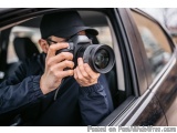 The image shows a man sitting in the driver's seat of a car, holding a DSLR camera and taking a photo. He is wearing a black...