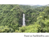 The image shows a large waterfall cascading down a steep hillside covered in lush greenery. The waterfall is located in the c...