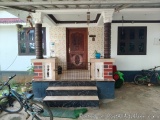 The image shows the entrance of a small house with a wooden door and two windows. The house has a white exterior with a red t...