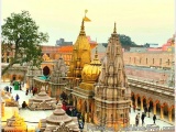 The image is a photograph of a Hindu temple complex. The temple is located in a city with other buildings and structures visi...