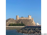 The image shows a castle perched on a rocky cliff overlooking the ocean. The castle is made of stone and has multiple towers...