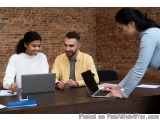 The image shows three people sitting at a wooden table with laptops in front of them. The table is in an office with a brick...