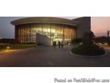 The image shows a modern building with a curved roof and large windows. The building is located in a park-like setting with a...