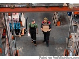 The image shows two men in a warehouse, standing in front of a large warehouse rack. They are both wearing overalls and hats,...