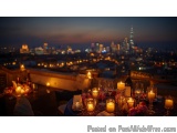 The image shows a table set up on a rooftop terrace with a beautiful view of the city at night. The table is set with a blue...