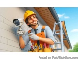 The image shows a young man wearing a yellow hard hat and overalls, holding a security camera in his hand. He is standing in...