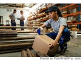 The image shows a young woman in a warehouse, wearing a blue cap and a gray t-shirt, kneeling on the floor and holding a card...