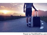 The image shows a person walking on a road with a suitcase. The person is wearing a blue t-shirt, shorts, and sneakers. The s...