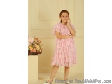 The image shows a young girl standing in front of a beige wall with a vase of flowers on the left side. She is wearing a pink...