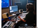 The image shows a young boy sitting at a desk in a recording studio. He is wearing a green t-shirt and has curly hair. The de...