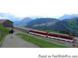 The image shows a red and white train on a track in a mountainous area. The train is stopped at a station with a wooden build...