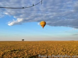 The image shows a hot air balloon flying over a vast open field. The balloon is yellow and red striped and is in the center o...