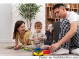 The image shows a young family of three - a man, a woman and a baby - playing with colorful toys on the floor in a living roo...