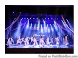 The image is a photograph of a stage with a large group of dancers performing on it. The stage is lit up with blue and purple...
