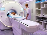 The image shows a medical examination room with a large CT scan machine. The machine is white and has multiple screens on the...