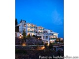 The image shows a large, luxurious mansion on a hillside at night. The mansion is white with multiple levels and balconies, a...