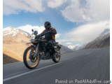 The image shows a person riding a motorcycle on a winding mountain road. The rider is wearing a black helmet, an orange jacke...