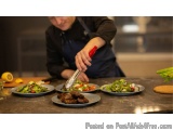 The image shows a chef in a kitchen preparing a meal. He is wearing a black apron and is standing behind a countertop with th...