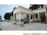 The image shows a large white building with a gazebo-like structure in the center. The building has multiple columns and a pe...