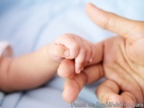 The image shows a close-up of a baby's hand being held by a person's hand. The hand is gently cradling the baby's fingers, wi...