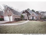 The image shows a single-story brick house with a sloping roof and a garage. The house is surrounded by a well-manicured lawn...