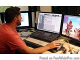 The image shows a man sitting at a desk in front of two computer monitors. He is wearing a red t-shirt and appears to be work...