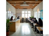 The image shows a classroom with a high ceiling and wooden beams. The room is filled with desks and chairs arranged in rows,...