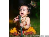 The image is a close-up portrait of a young Indian child, likely a Hindu deity, sitting on the grass. The child is wearing a...