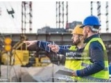 The image shows two construction workers wearing hard hats and high visibility vests on a construction site. They are standin...