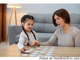 The image shows a mother and daughter playing a board game together at a wooden table. The mother is sitting on the right sid...