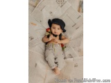 The image is a photograph of a baby lying on a bed of newspaper. The baby is wearing a black hat and is holding a red rose in...
