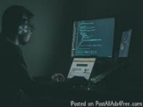 The image shows a person sitting at a desk in front of a computer monitor. The person is wearing a black hoodie and has a fac...