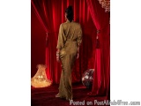 The image shows a woman standing in front of a red curtain. She is wearing a long, flowing, olive green dress with a high nec...