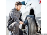 In this image, we can see a man working on a car tire. He is kneeling on the ground and is holding the tire with both hands....