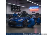 The image shows a blue Nissan GT-R sports car in a workshop. There are three men working on the car, two of them are kneeling...