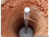 The image shows a small hole in the ground with a metal pole in the center. The pole is cylindrical in shape and appears to b...