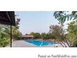 The image shows a large swimming pool with crystal clear blue water. The pool is surrounded by lush greenery and trees, and t...