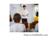 The image shows a young man standing in front of a group of people in a classroom. He is wearing a white sweater and jeans an...