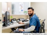 The image shows a man sitting at a desk in an office. He is wearing a blue shirt and has a beard and is smiling at the camera...