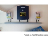 The image shows a blue-colored solar panel mounted on a white wall. The panel has a digital display screen and two wires conn...