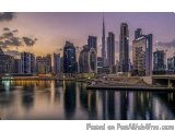 The image is a panoramic view of a city skyline at dusk. The sky is a deep purple color with the sun setting in the backgroun...