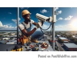 The image shows a construction worker wearing a hard hat and overalls, working on a security camera mounted on a pole. He is...