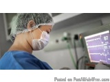 The image shows a person wearing a surgical mask and a hairnet, working in an operating room. The person is wearing blue scru...
