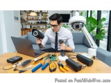 The image shows a young man sitting at a desk with a laptop and various tools and equipment scattered around him. He is weari...