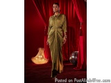 The image shows a woman standing in front of a red curtain. She is wearing a long, flowing, olive green dress with a belt cin...