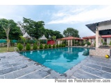 The image shows a large swimming pool with crystal clear blue water. The pool is surrounded by a stone patio with a few loung...