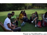 The image shows a group of six people sitting on a grassy lawn in a park. They are all wearing casual clothes and have backpa...